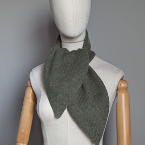 May include: A knitted olive green scarf displayed on a mannequin. The scarf is made of a ribbed knit fabric and has a unique shape, with pointed ends. The scarf is draped around the mannequin's neck, showcasing its design and texture.