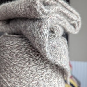 Puede incluir: Primer plano de una madeja de hilo gris con un trozo doblado del mismo hilo encima. El hilo tiene un aspecto texturizado con motas de color. El trozo doblado muestra el patr&oacute;n de punto. El hilo es probablemente para proyectos de tejido o ganchillo.