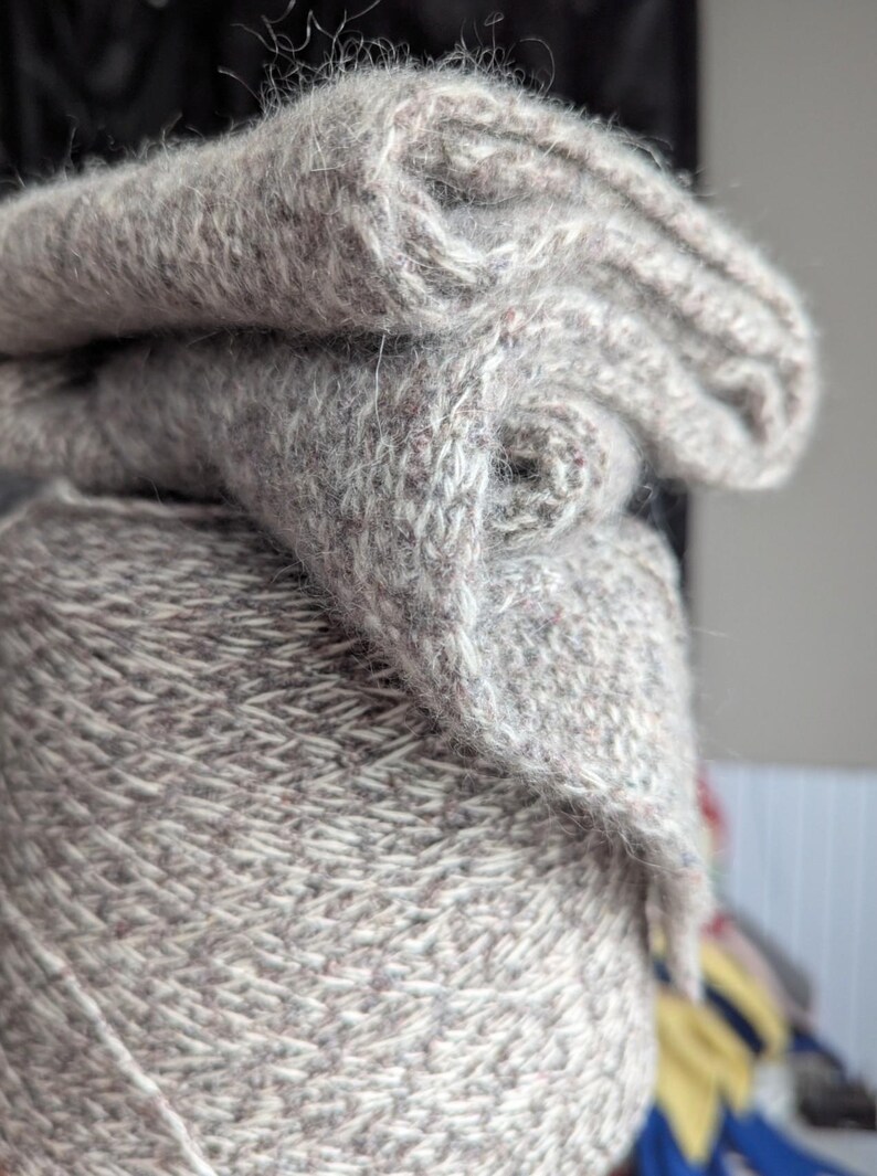 Puede incluir: Primer plano de una madeja de hilo gris con un trozo doblado del mismo hilo encima. El hilo tiene un aspecto texturizado con motas de color. El trozo doblado muestra el patr&oacute;n de punto. El hilo es probablemente para proyectos de tejido o ganchillo.