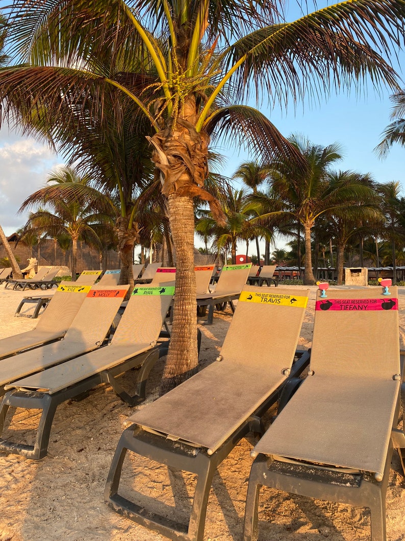 May include: Beach scene with several lounge chairs on the sand, each with a reserved sign. Palm trees are in the background under a blue sky. The signs have names like Travis and Tiffany.
