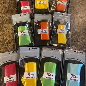 May include: A collection of colorful chair savers in clear plastic bags. Each bag contains a folded ribbon in various colors, including red, yellow, orange, and green. Each bag has a label that reads "CHAIR SAVER".