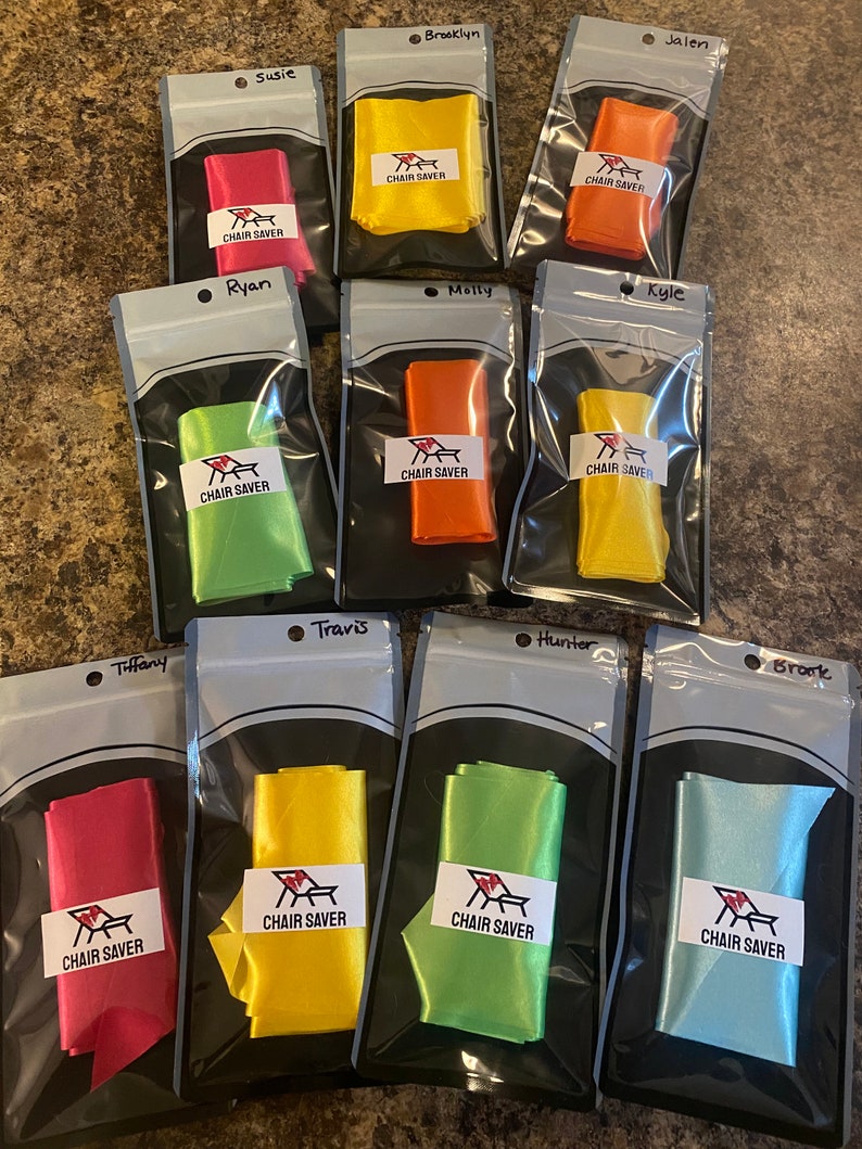 May include: A collection of colorful chair savers in clear plastic bags. Each bag contains a folded ribbon in various colors, including red, yellow, orange, and green. Each bag has a label that reads "CHAIR SAVER".