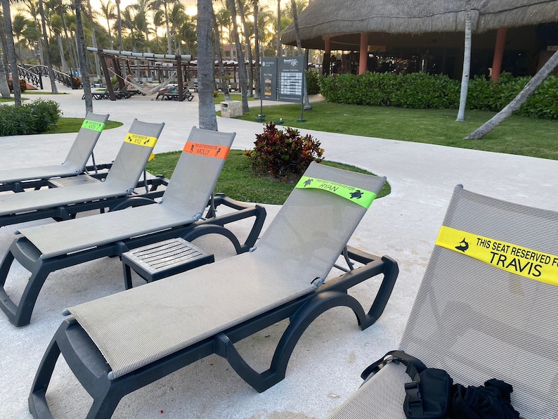 May include: Several gray lounge chairs with black frames are arranged outdoors. Each chair has a reserved seat band with names like "Travis", "Molly", and "Ryan". The scene is set near a pool or beach area, with palm trees in the background.