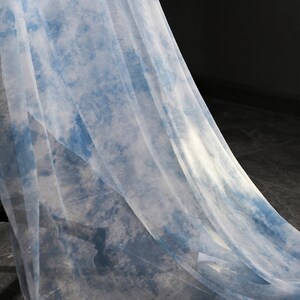 May include: A sheer, light blue fabric with a tie-dye pattern. The fabric is draped over a dark surface.