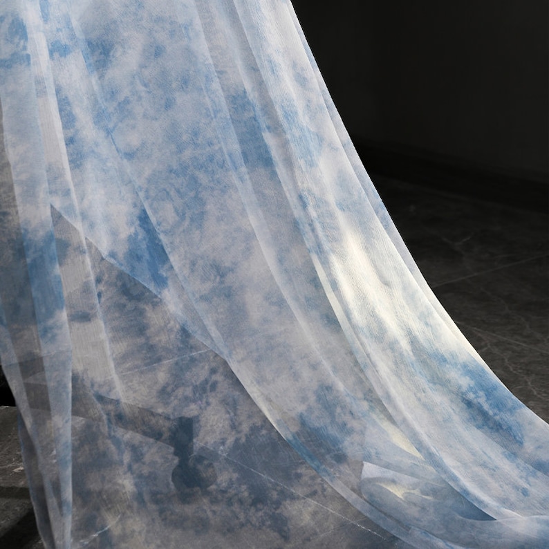 May include: A sheer, light blue fabric with a tie-dye pattern. The fabric is draped over a dark surface.