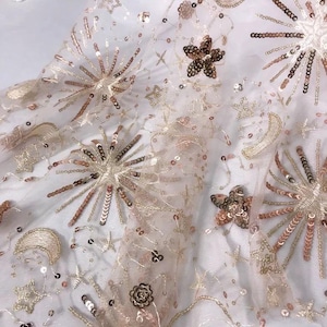 May include: A delicate, sheer fabric with a subtle pink hue, adorned with gold and rose gold sequins in the shape of stars, moons, and flowers. The sequins are arranged in a repeating pattern, creating a celestial-inspired design.
