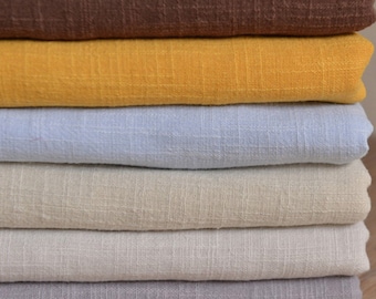 May include: A stack of six folded fabric pieces featuring a visible grid-like texture. The colors from top to bottom are dark brown, mustard yellow, light blue, cream, off-white, and light gray. Each piece of fabric has a consistent weave.