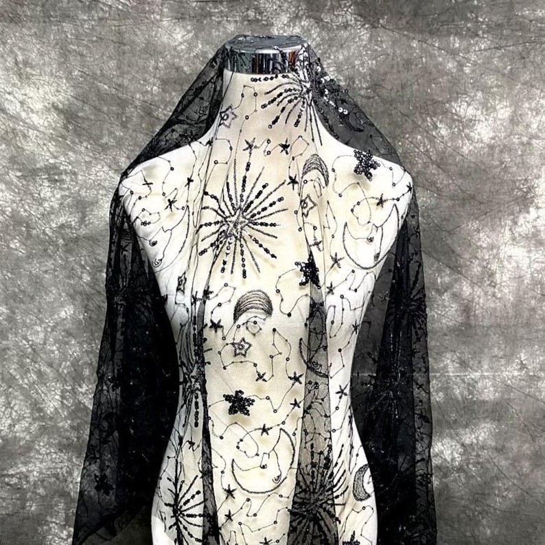 May include: Black sheer fabric with a celestial design featuring stars, moons, and constellations. The fabric is embellished with sequins and beads.