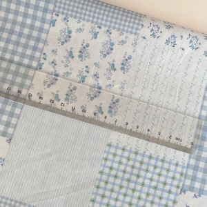 May include: A patchwork fabric with various patterns, including blue gingham, floral prints, and striped sections. A clear ruler with centimeter markings is laid across the fabric. The fabric's color palette is primarily light blue and white.