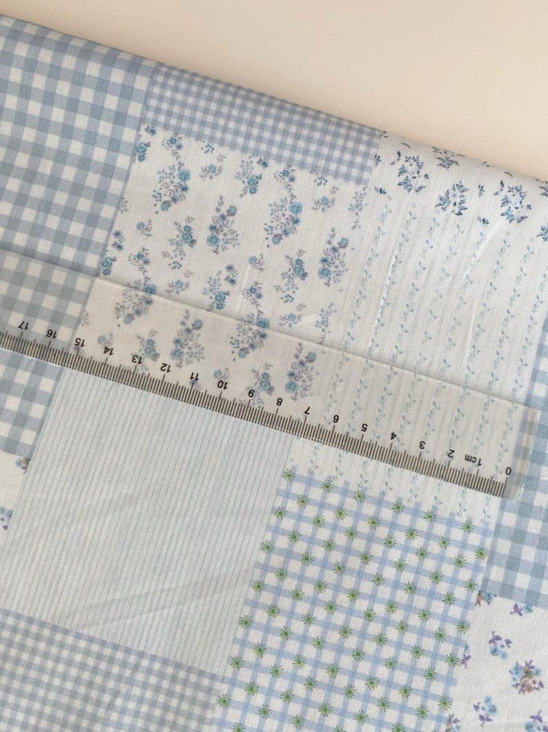 May include: A patchwork fabric with various patterns, including blue gingham, floral prints, and striped sections. A clear ruler with centimeter markings is laid across the fabric. The fabric's color palette is primarily light blue and white.