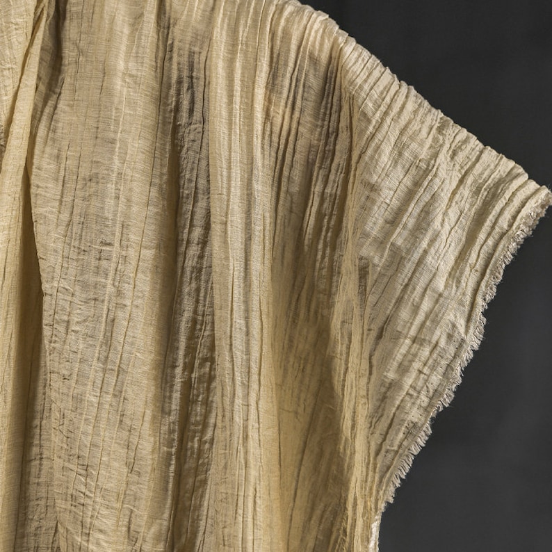 Pu&ograve; includere: Un tessuto beige chiaro semi-trasparente con una distinta consistenza stropicciata verticale. Il materiale presenta un drappeggio sciolto e fluido e un bordo grezzo sfilacciato sul lato, mostrando una trama leggera con sottili variazioni.