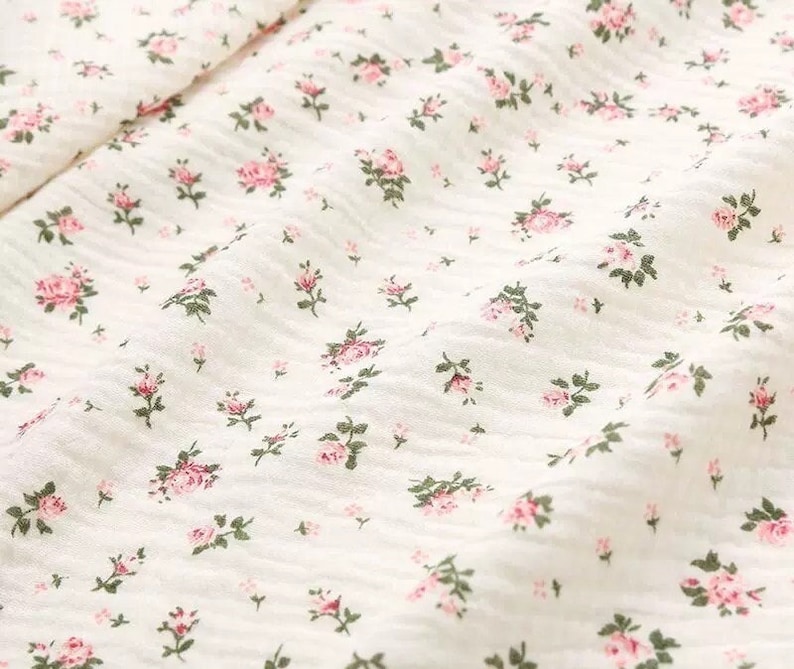 Op de afbeelding: Witte stof met een klein roze bloemenpatroon. De stof heeft een licht getextureerd oppervlak.