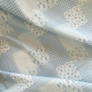 May include: A patchwork quilt featuring a mix of patterns in shades of blue and white. The design includes gingham, floral, and striped sections, creating a textured and visually appealing textile. The quilt is folded, showing the fabric's drape.