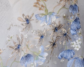 以下が含まれることがあります： 青とベージュの色合いの刺繍された花柄が施された透き通った生地。生地には、小さなデイジーや、より大きく手の込んだ花など、さまざまな花の形が施されています。