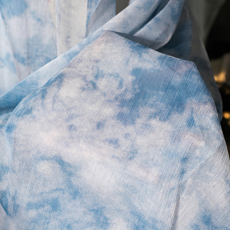 May include: A close-up of a blue and white tie-dye fabric. The fabric has a soft, flowing texture and a subtle sheen.