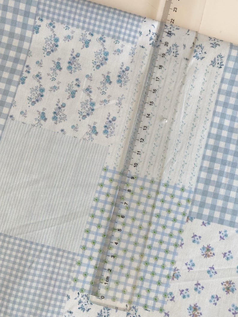 May include: A patchwork fabric with various patterns, including blue gingham, floral prints, and striped sections. A ruler is placed on the fabric, showing measurements up to 23 cm.