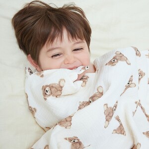 May include: A young child is wrapped in a white muslin blanket with a brown teddy bear print. The child is smiling and looking at the camera.