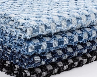 May include: A stack of four folded fabric pieces featuring a checkered pattern with frayed, raw edges. The colours transition from light blue to medium blue, dark blue, and black, each paired with white in a repeating square grid design.