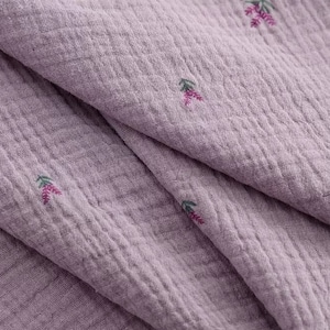 Op de afbeelding: Close-up van een zachte, textuurrijke, lavendelkleurige stof met een subtiel bloemenpatroon. De stof is dubbelgevouwen, waardoor het patroon aan beide kanten zichtbaar is.