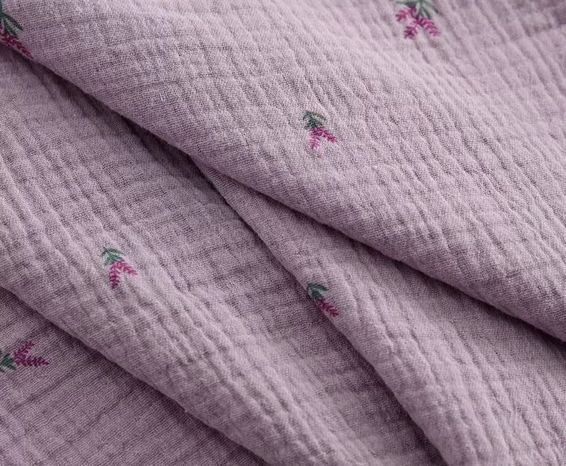 Op de afbeelding: Close-up van een zachte, textuurrijke, lavendelkleurige stof met een subtiel bloemenpatroon. De stof is dubbelgevouwen, waardoor het patroon aan beide kanten zichtbaar is.