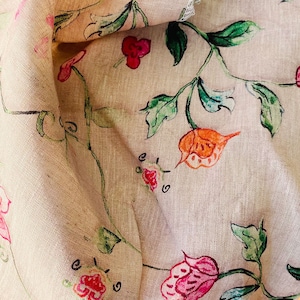 May include: A light beige fabric featuring a hand-drawn floral pattern with green vines, leaves, and flowers in shades of pink, red, and orange. The textile has a visible woven texture and folds that create depth across the surface of the material.