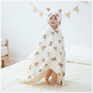 May include: A white hooded towel with a brown bear print. The towel has a hood with bear ears and a button closure. The towel is made of soft, absorbent material.