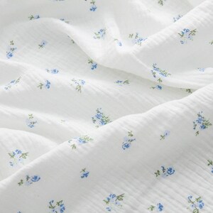 Op de afbeelding: Witte stof met een delicaat blauw bloemenpatroon. De stof heeft een licht getextureerd oppervlak.