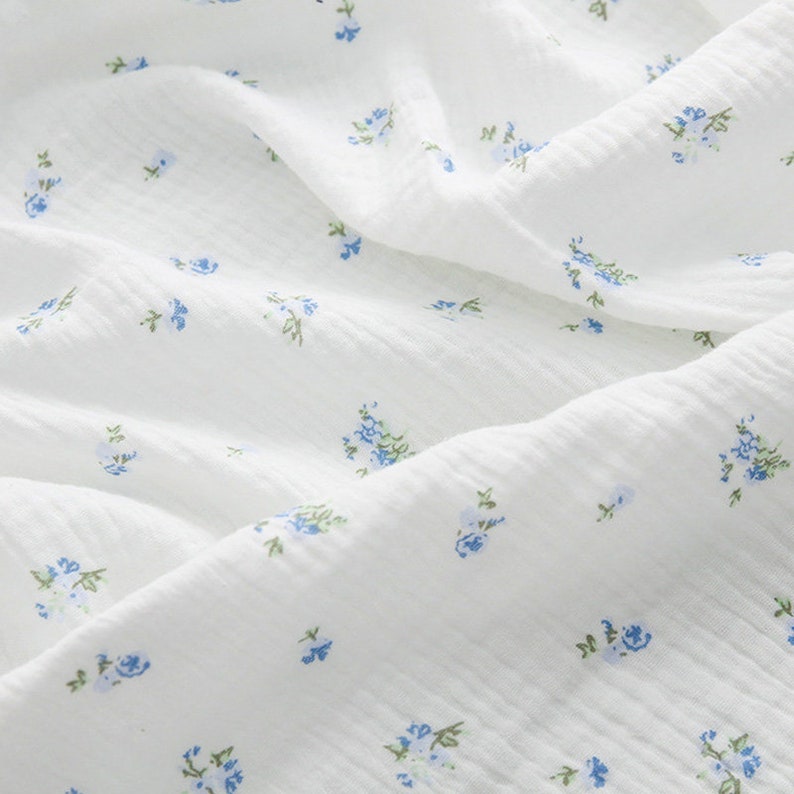 Op de afbeelding: Witte stof met een delicaat blauw bloemenpatroon. De stof heeft een licht getextureerd oppervlak.