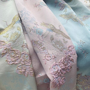 Könnte beinhalten: Nahaufnahme eines Stoffes mit einem rosa-blauen Blumenmuster. Der Stoff zeigt ein sich wiederholendes Muster aus Kranichen, Blumen und Blättern. Der Stoff hat einen dezenten Schimmer.