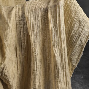 Pu&ograve; includere: Un tessuto beige chiaro con una distinta consistenza stropicciata verticale. Il materiale presenta una serie di creste parallele in rilievo che corrono lungo la sua lunghezza, creando un aspetto plissettato. I bordi del tessuto mostrano una finitura leggermente sfilacciata.