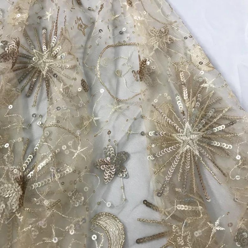 May include: Beige tulle fabric with gold embroidered stars and sequins. The fabric has a delicate, airy feel and is perfect for adding a touch of sparkle to any project.