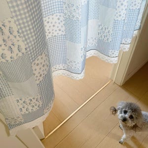 May include: Light blue and white patchwork curtain with floral and gingham patterns. The curtain has a white lace trim along the bottom edge. A small, gray dog is in the foreground.