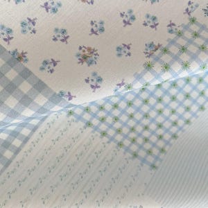 May include: Close-up of a fabric with a floral and gingham pattern. The fabric features small blue and purple flowers on a white background, along with light blue gingham and striped sections. The overall aesthetic is soft and delicate.