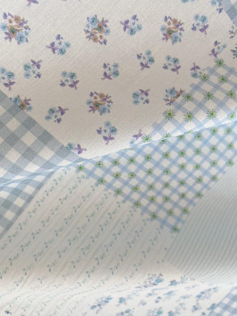 May include: Close-up of a fabric with a floral and gingham pattern. The fabric features small blue and purple flowers on a white background, along with light blue gingham and striped sections. The overall aesthetic is soft and delicate.