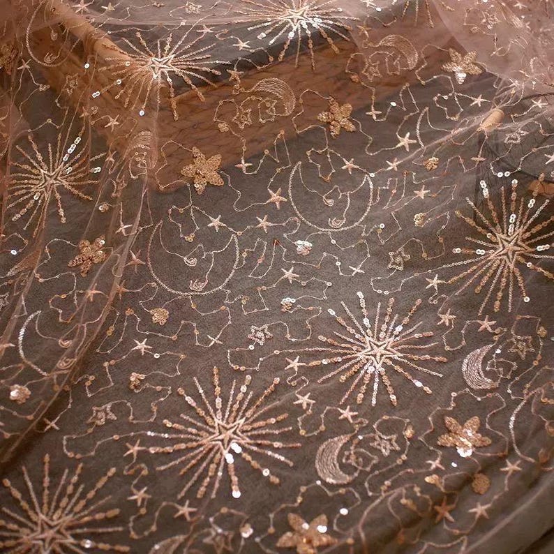 May include: A sheer pink fabric with a delicate pattern of stars, moons, and sequins. The fabric has a subtle shimmer and is perfect for crafting or adding a touch of sparkle to any project.