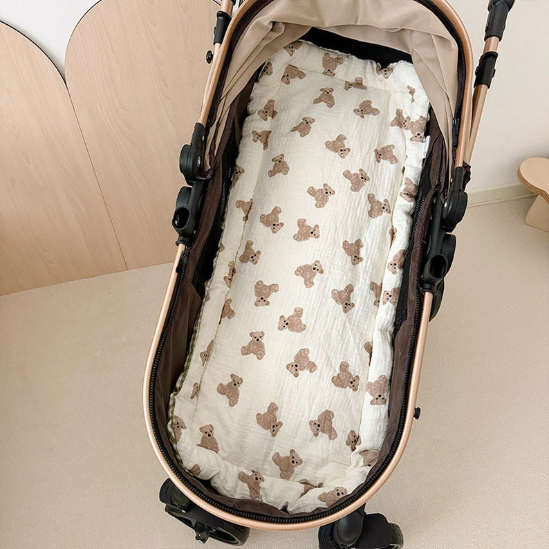 May include: A white stroller liner with a brown teddy bear print. The liner is in a stroller with brown fabric and black wheels.
