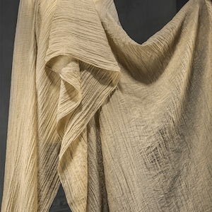 Pu&ograve; includere: Un tessuto beige con una superficie increspata e strutturata. Il materiale &egrave; drappeggiato in pieghe larghe e fluide, creando ombre e riflessi profondi sulla superficie irregolare e ondulata del tessuto, mettendone in risalto la leggerezza.