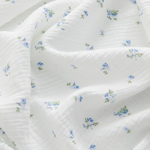 Op de afbeelding: Witte stof met een klein blauw bloemenpatroon. De stof heeft een textuur en is licht gekreukt.