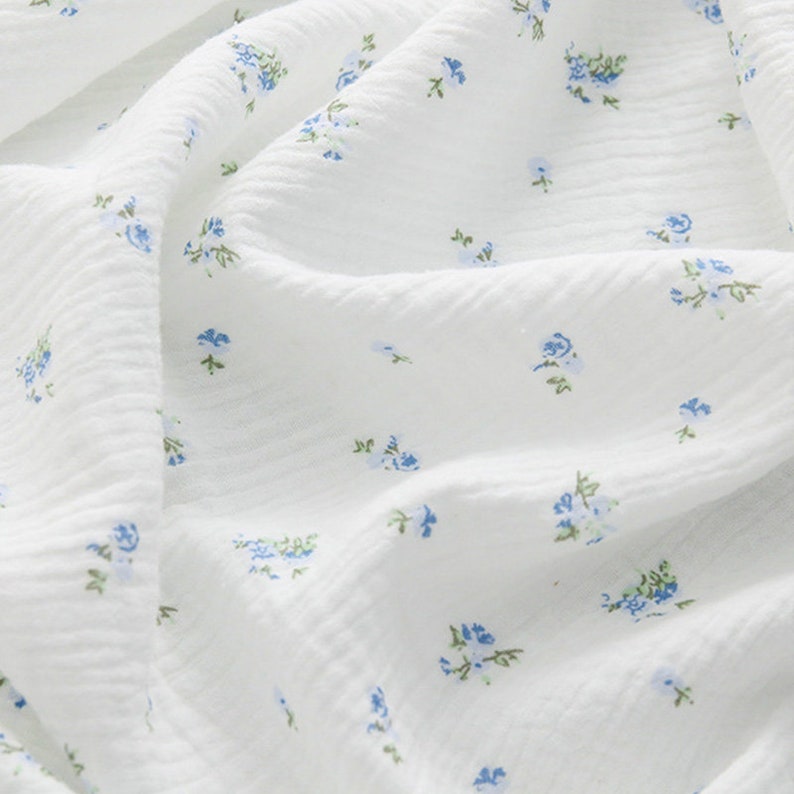 Op de afbeelding: Witte stof met een klein blauw bloemenpatroon. De stof heeft een textuur en is licht gekreukt.