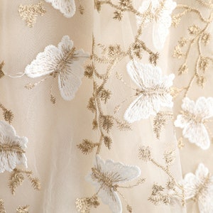 Puede incluir: Una tela beige transparente con intrincados bordados de mariposas blancas y enredaderas metálicas doradas. El delicado material de malla está adornado con motivos de mariposas y patrones de ramas florales en hilo dorado metálico.