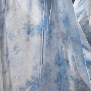 May include: Close-up of sheer fabric with a blue and white tie-dye pattern. The fabric is draped and folded, creating a soft and ethereal look.