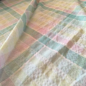 Peut inclure: Un tissu textile présentant un motif à carreaux doux dans des tons pastel de rose, vert et jaune. Le matériau affiche un design à grille subtil avec des lignes fines et entrecroisées formant des carrés sur toute la surface du tissu.