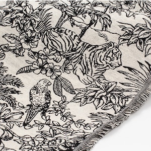 May include: A woven textile featuring a black and cream toile-style pattern of a tiger resting among tropical foliage and a perched parrot. The fabric has a textured surface and a finished edge with a short, delicate fringe along the side.