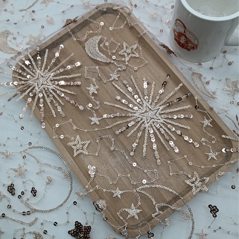 May include: A close-up of a piece of fabric with a delicate, white mesh background and silver sequins arranged in the shape of stars and a crescent moon.