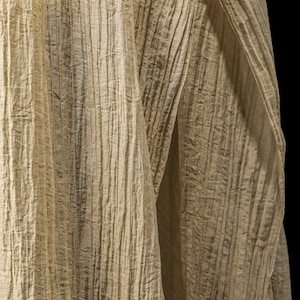 Pu&ograve; includere: Un tessuto beige chiaro con una distinta consistenza stropicciata verticale. Il materiale presenta profonde pieghe parallele che creano una serie di creste e valli su tutta la superficie, risultando in un aspetto altamente strutturato.