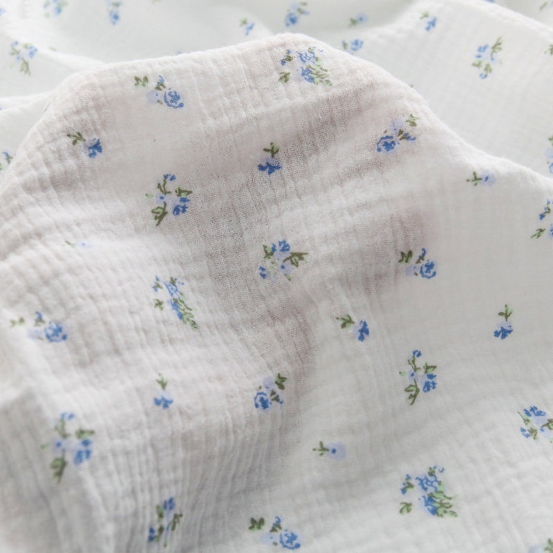 Op de afbeelding: Witte stof met een klein blauw bloemenpatroon. De stof heeft een textuur en is licht gekreukt.