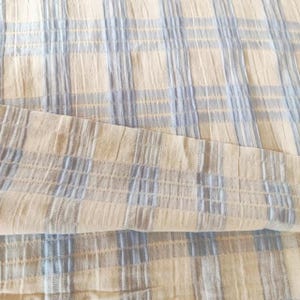 Può includere: Un pezzo di tessuto con un motivo a quadri color crema e azzurro. Il materiale presenta una distinta consistenza stropicciata e increspata su tutta la superficie, creando un design a griglia con strisce verticali e orizzontali.