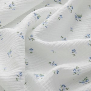 Op de afbeelding: Witte stof met een klein blauw bloemenpatroon. De stof heeft een textuur, licht gekreukeld.