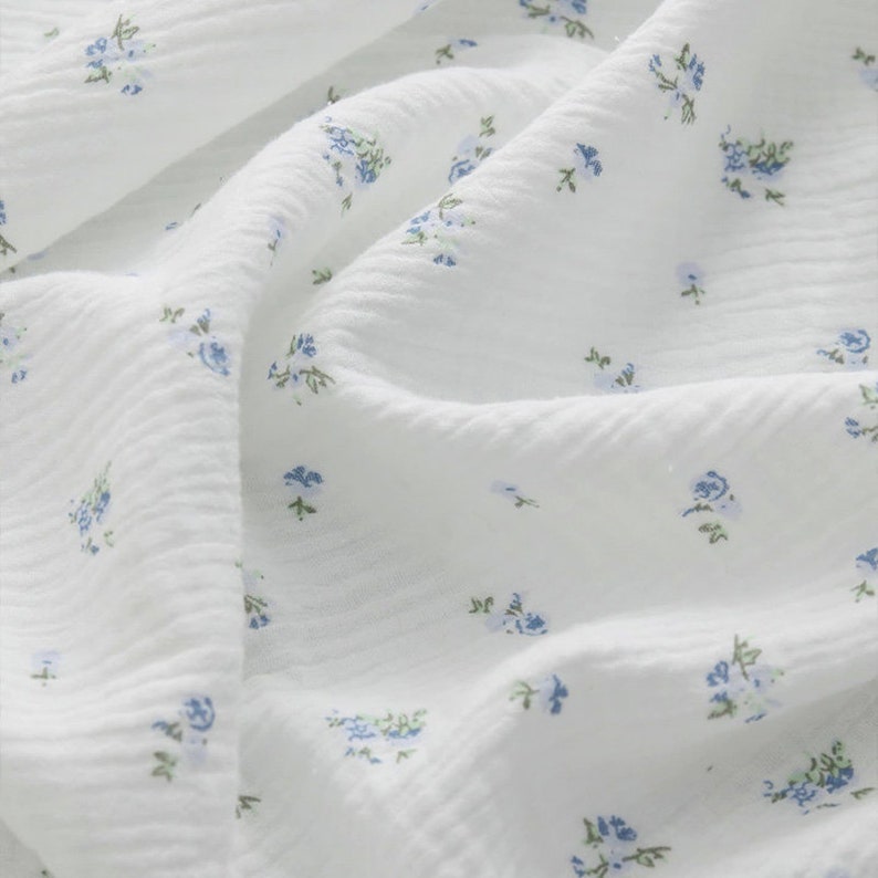 Op de afbeelding: Witte stof met een klein blauw bloemenpatroon. De stof heeft een textuur, licht gekreukeld.