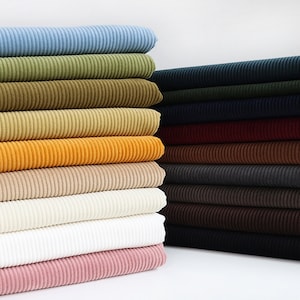 May include: A collection of folded corduroy fabric stacks arranged in two columns. The left stack features light blue, olive green, mustard, beige, white, and pink hues. The right stack displays dark navy, forest green, burgundy, brown, and black.